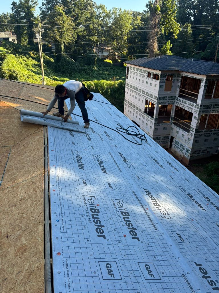 Roofers installing GAF underlayment on new roof with wood decking, surrounded by greenery and a residential construction site