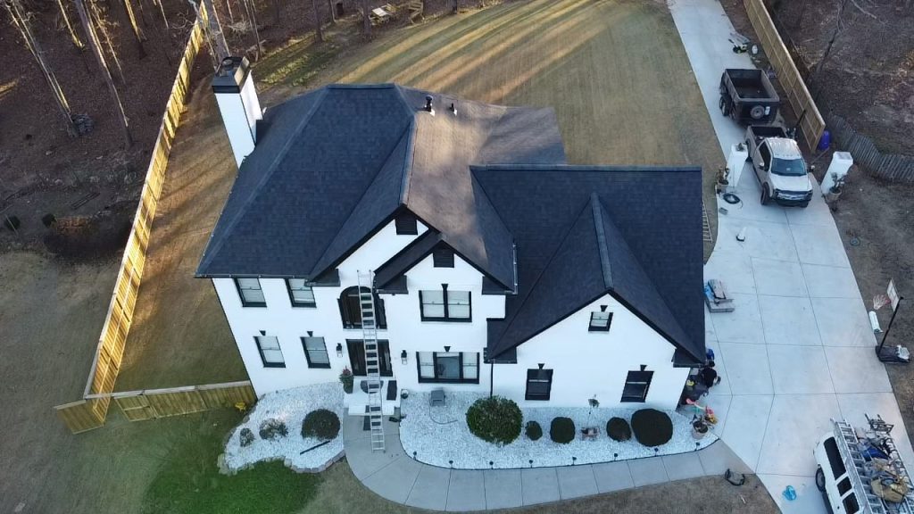 White home with black roof, crew installing shingles, HD Pro Roofing equipment and truck in driveway.