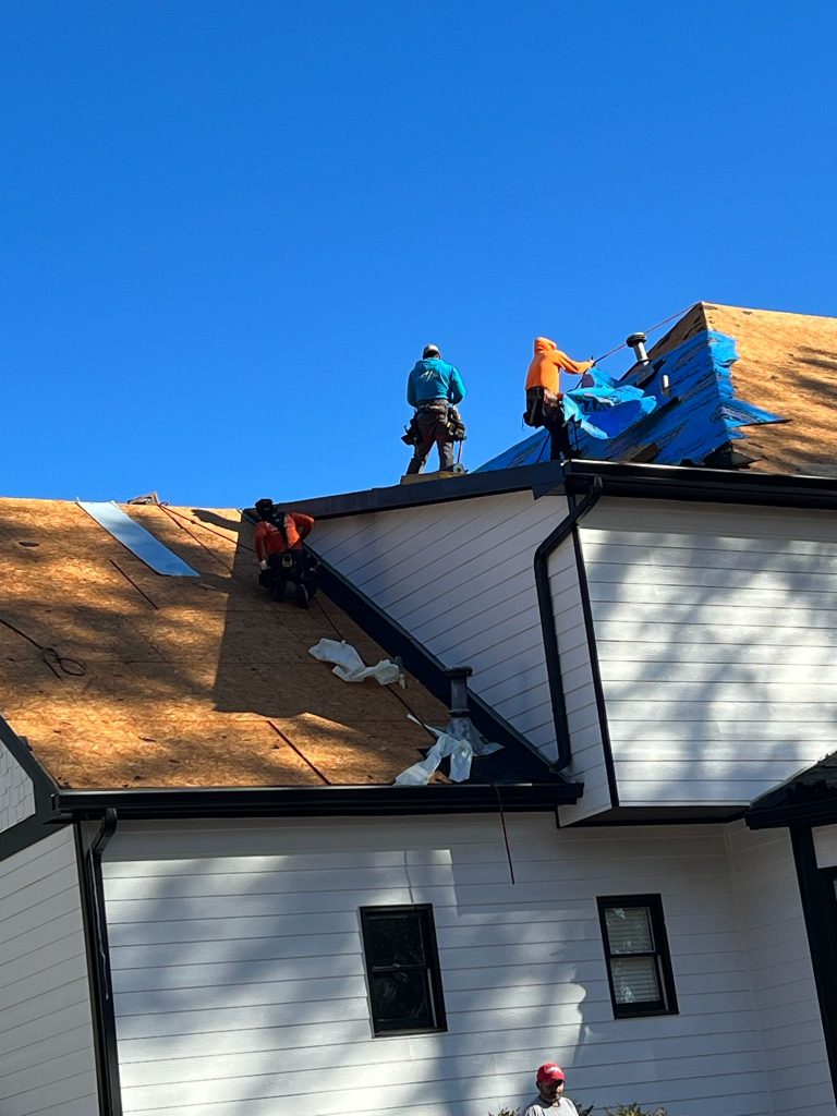 Roofers installing new shingles and tarps on white house roof under clear blue sky during residential roofing project.