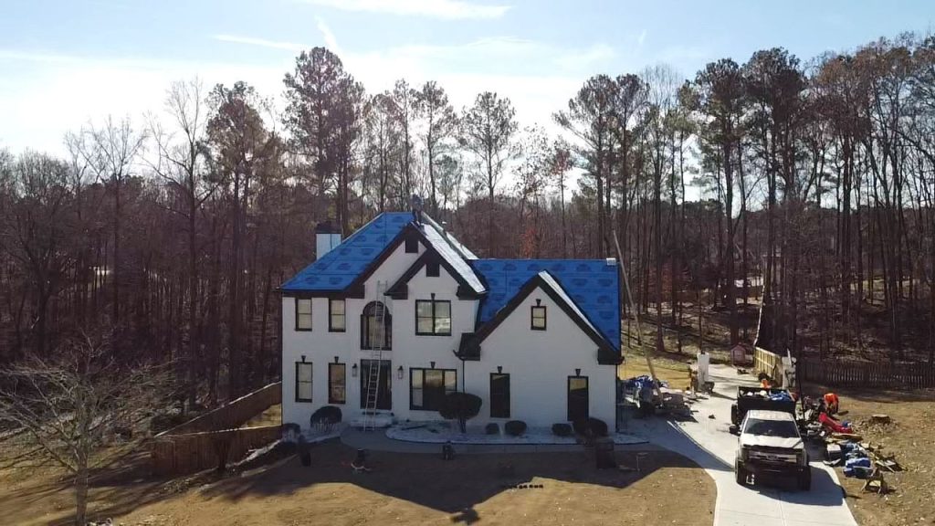 White two-story house with new blue shingle roofing, surrounded by trees, roofing materials, and HD Pro Roofing truck in driveway.