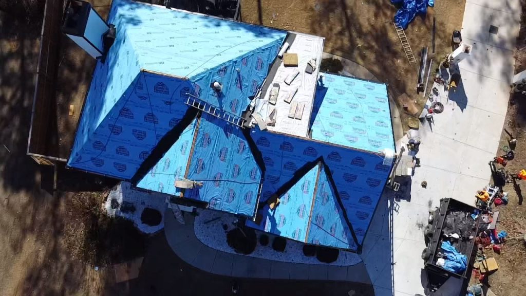 Aerial view of roofing project with blue synthetic underlayment, workers on ladders, and HD Pro Roofing equipment on site.