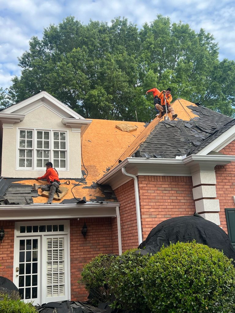 Roofing contractors in orange shirts repairing a red brick home roof, tearing off old shingles under blue sky for HD Pro Roofing.