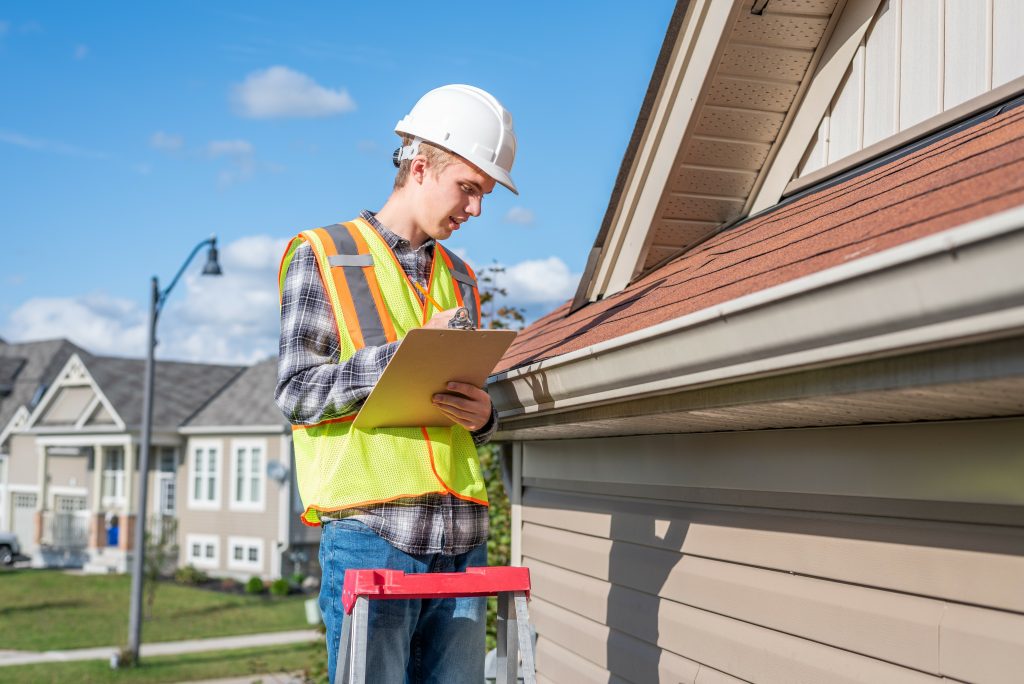 Roof inspector in safety gear examining shingles on a residential roof using a clipboard under a sunny sky.