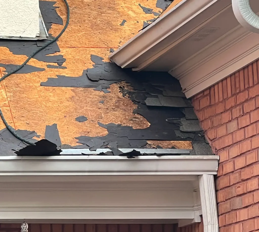 Damaged asphalt shingles and exposed plywood roof deck on red brick house highlighting roof deterioration for HD Pro Roofing repairs.